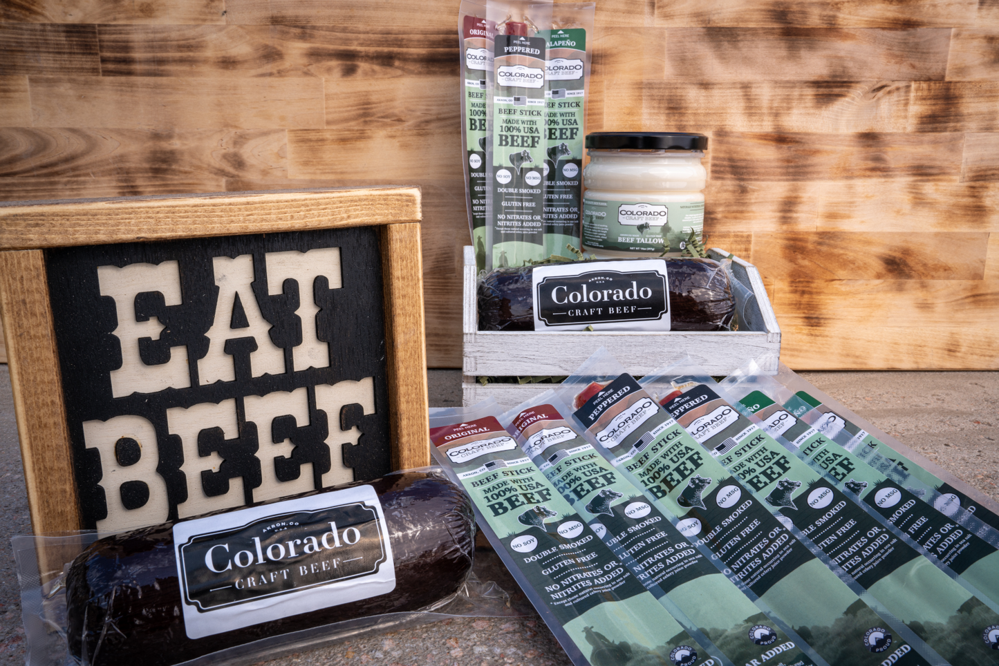 A gift box of beef products including beef sticks, smoked jalapeno beef tallow, and summer sausage placed on a wooden surface with a sign that reads 'EAT BEEF'.